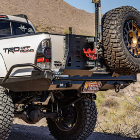 Silver 2nd Gen Toyota Tacoma TRD Off-Road in the desert with DRT swing out tire carrier closed, Hi-Lift jack and RotopaX mounted, bronze Method wheel spare tire